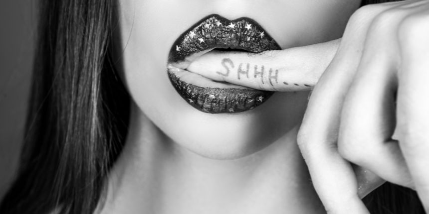 Sieviete ar spīdīgām lūpām maigi iekož pirkstā ar uzrakstu "SHHH", radot noslēpumainu un pavedinošu noskaņu.